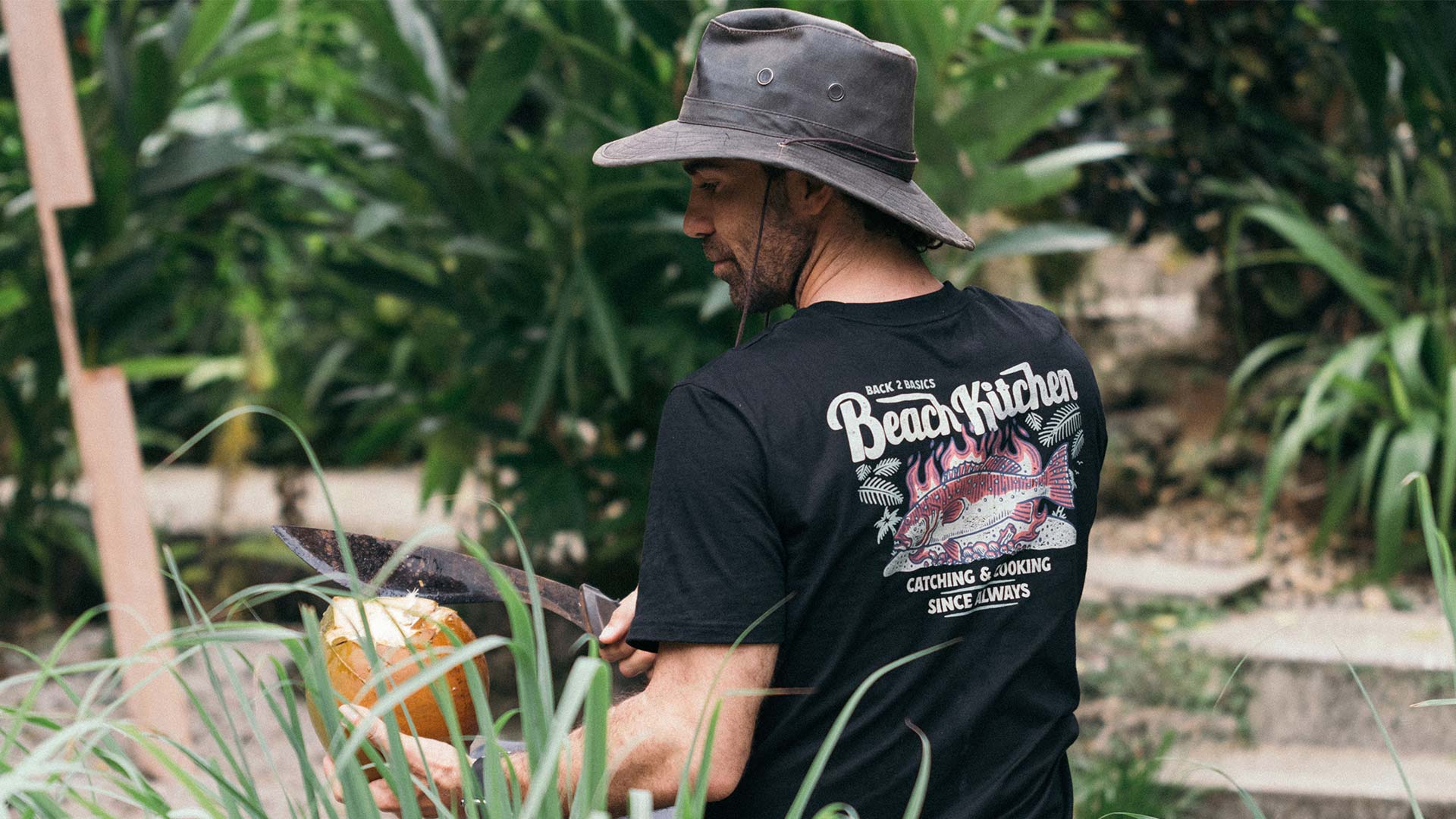 aaron from b2b adventures cutting a coconut in the bush while wearing a black back 2 basics beach kitchen graphic tee shirt