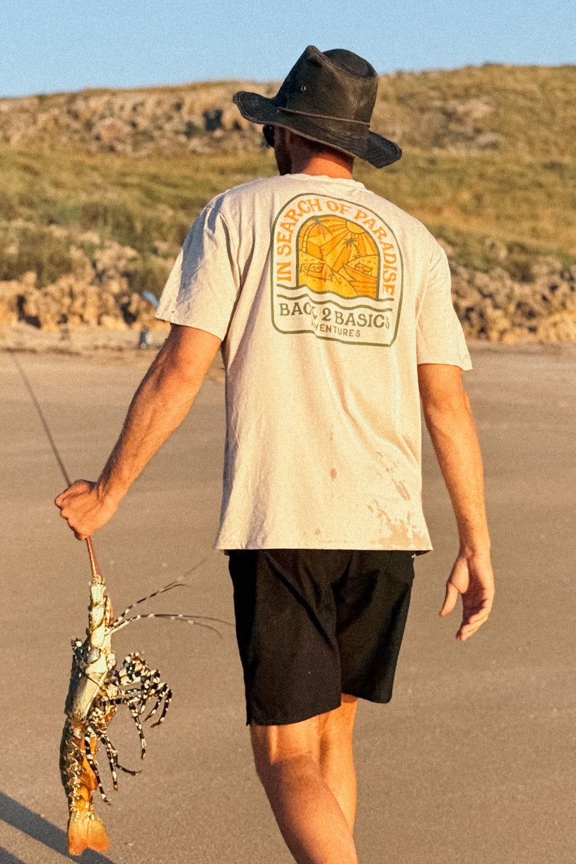 a man in a beige shirt with the words in search of paradise written on the back holding a large lobster or crayfish