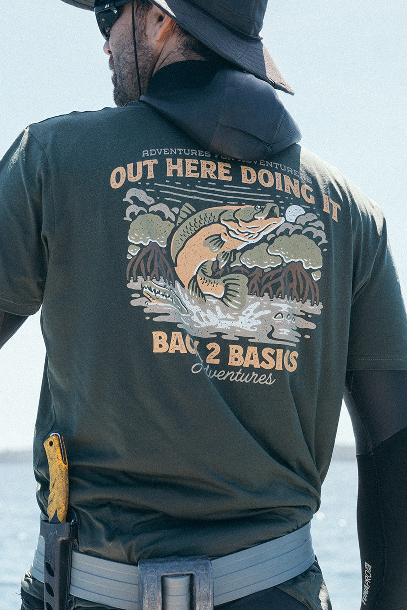 man standing in a boat with a spear gun about to go fishing wearing a forest green tee shirt that says back 2 basics adventures